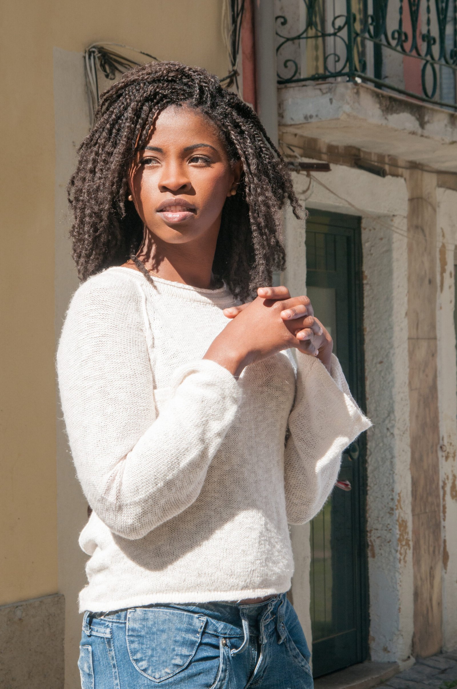 Pensive pretty young black woman standing outdoors. Woman keeping her hands clasped with old building in background. Tourism and leisure concept.